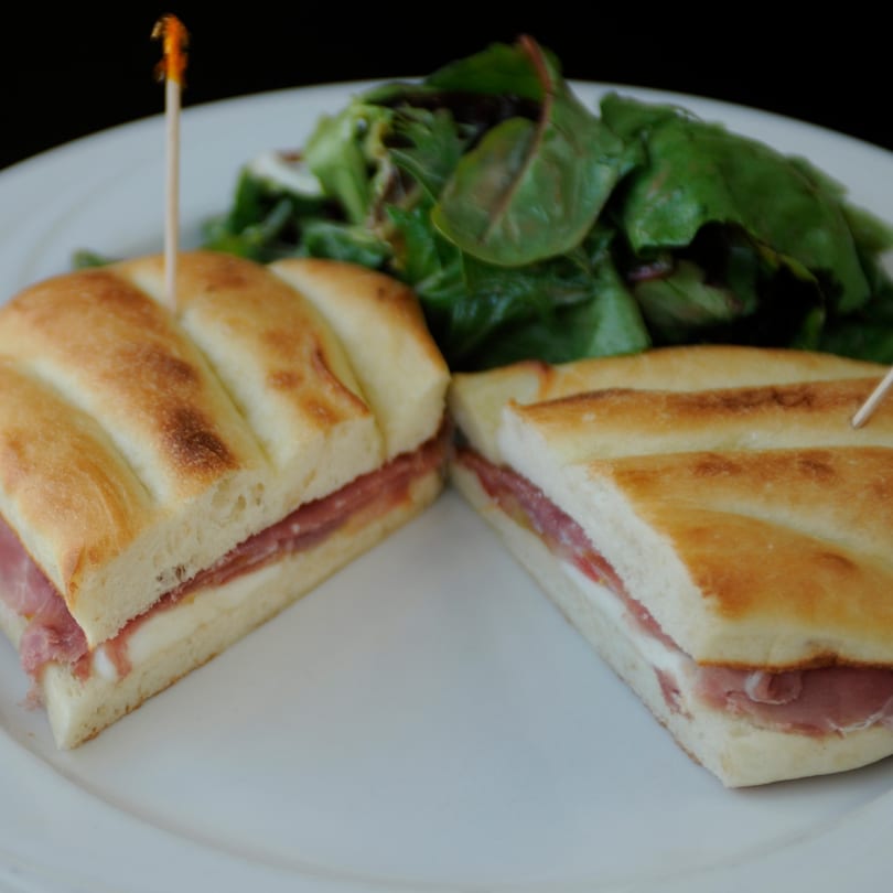 Two halves of a focaccia sandwich with prosciutto and mozzarella, served with a side salad.