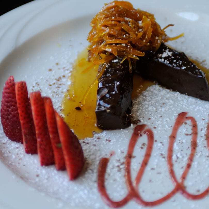 Chocolate dessert with candied orange peel, sliced strawberries, and raspberry drizzle on a white plate.