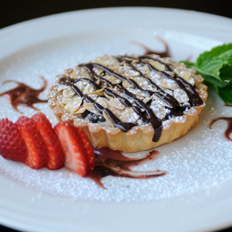 Almond and chocolate tart with powdered sugar, sliced strawberries, and mint on a white plate.