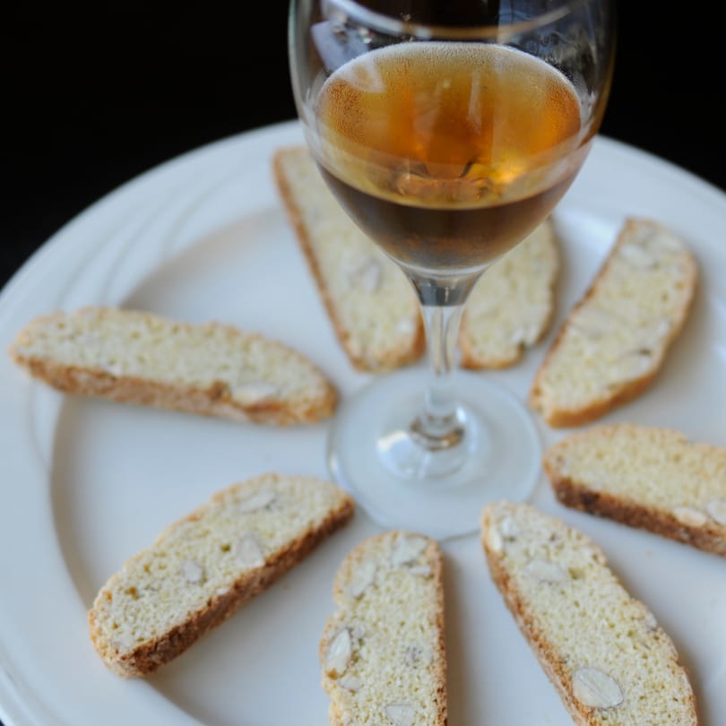 A glass of amber liquid surrounded by almond biscotti on a white plate at Breakfast At Vivoli Cafe.