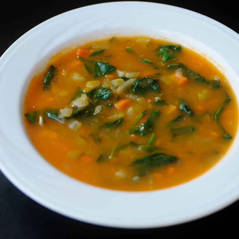 A white bowl filled with a vibrant orange vegetable soup, featuring spinach, carrots, and potatoes.