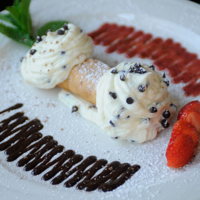 Two cannolis with chocolate chips and powdered sugar, garnished with strawberries and chocolate sauce.