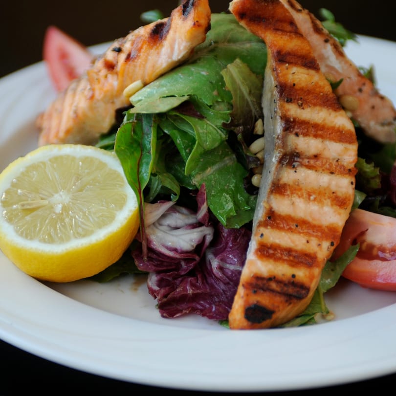 Grilled salmon fillets with a fresh green salad, tomato slices, and a lemon wedge on a white plate.